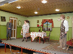 Tre skuespillere på en scene dekoreret som en stue med et spisebord og et falsk vindue med idyllisk udsigt over en nabogård. To af skuespillerne, den ene i en blomstret kjole med en brun håndtaske og en stråhat i hænderne, den anden i et blomstret forklæde og krøllet hår, kigger på den tredje, en mand som står ved døren med hænderne foldet klædt i grå bukser og jakke.