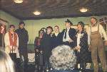 Otte skuespillere som står på scenen. To kigger op mod venstre, mens resten kigger ud mod publikum. Flere af dem ser ud til at være i gang med at synge.